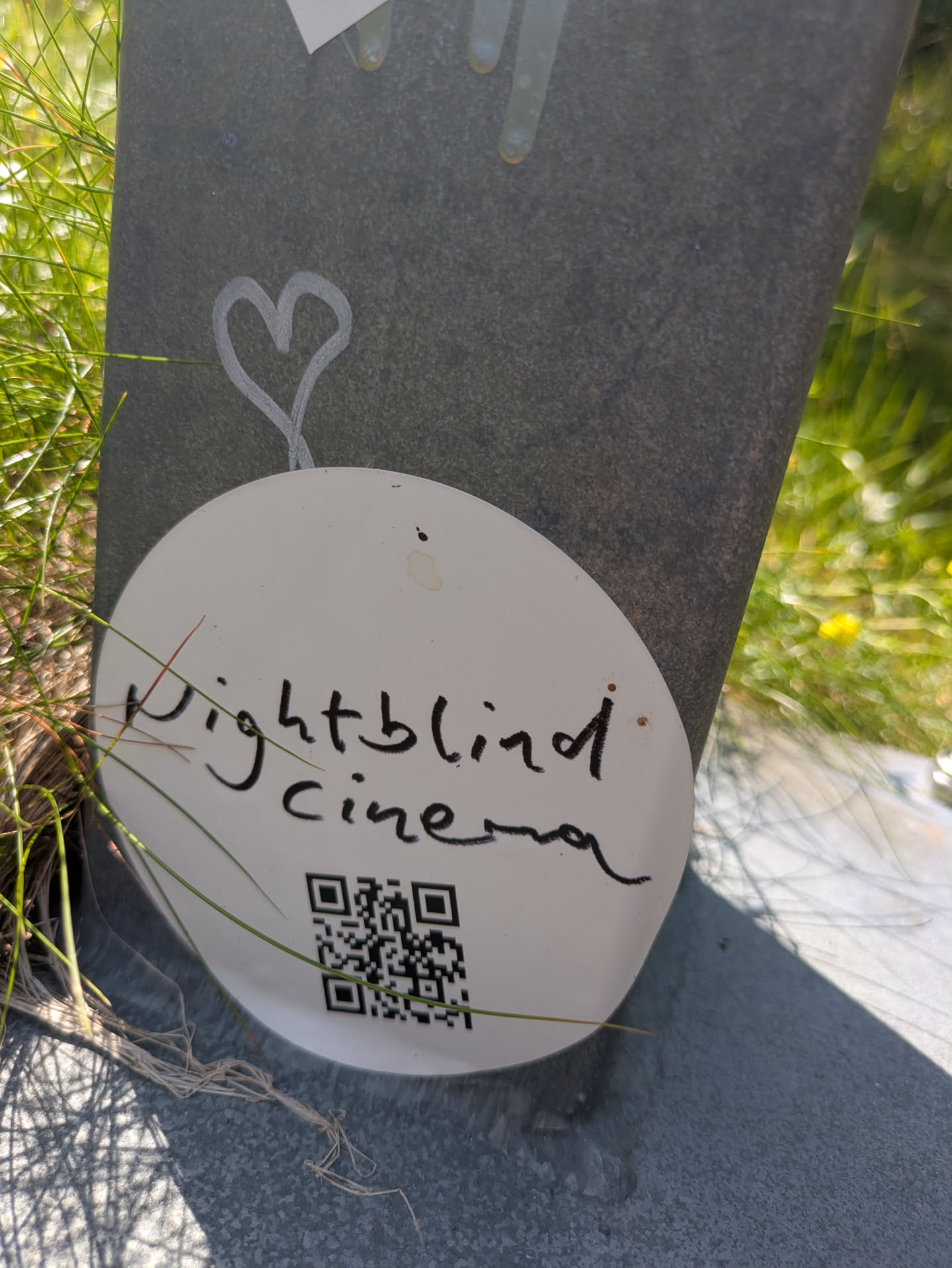 A white, circular sticker is affixed to a dark grey, textured surface, possibly concrete or stone. Handwritten in black cursive on the sticker are the words "Nightblind cinema". Below the text, a QR code is clearly visible. Above the sticker, on the grey surface, a simple white outline of a heart is drawn. The sticker is surrounded by green grass and plant material, suggesting an outdoor, possibly urban, environment.
