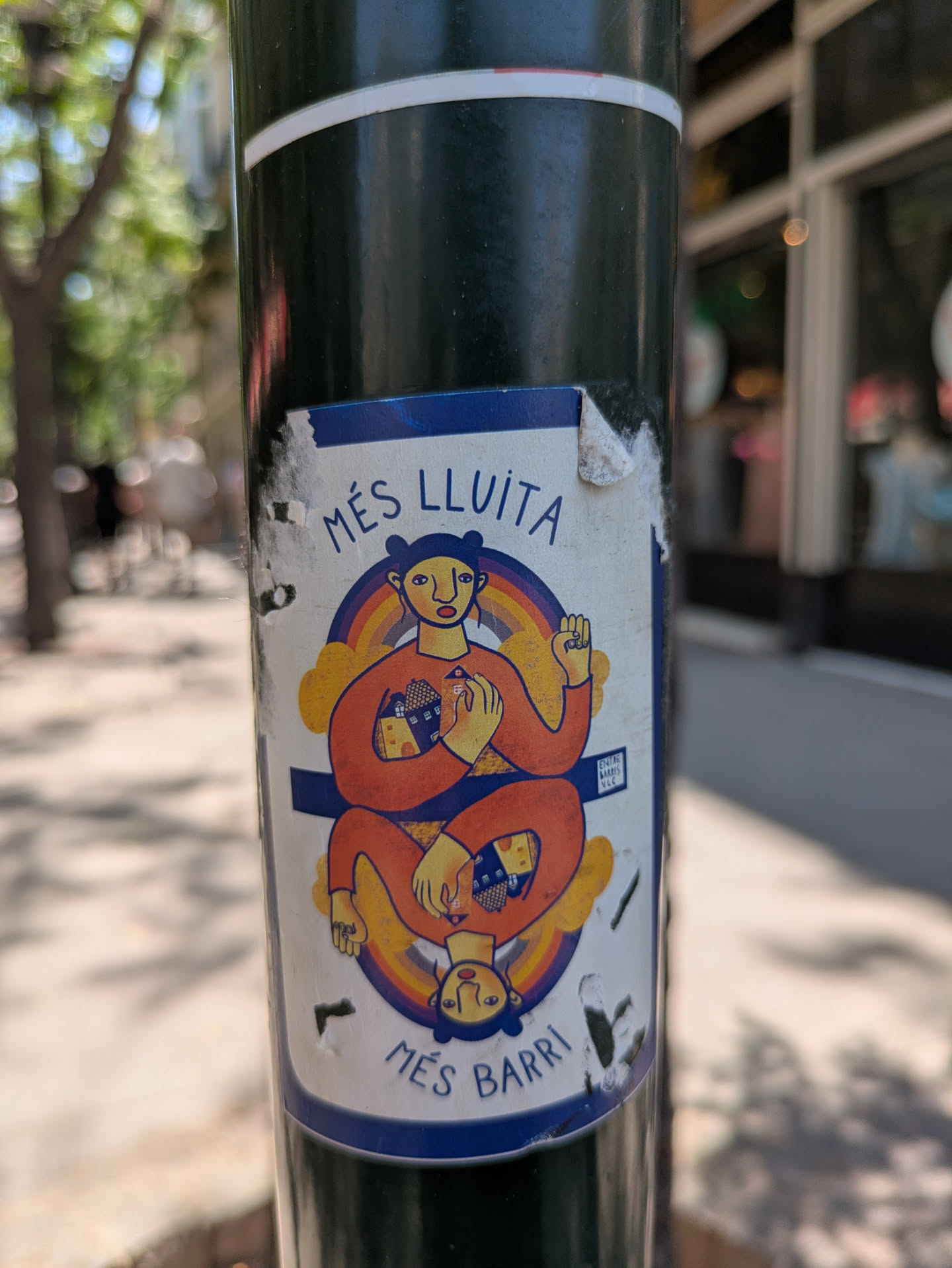 This sticker showcases a person in an orange outfit, sitting cross-legged, and mirrored with their upside-down reflection. They are holding a house in one hand and raising a fist in the other. The sticker is framed by a rainbow and clouds, adding a touch of whimsy. The text "Més Lluita Més Barri" is written on the top and bottom of the drawing.