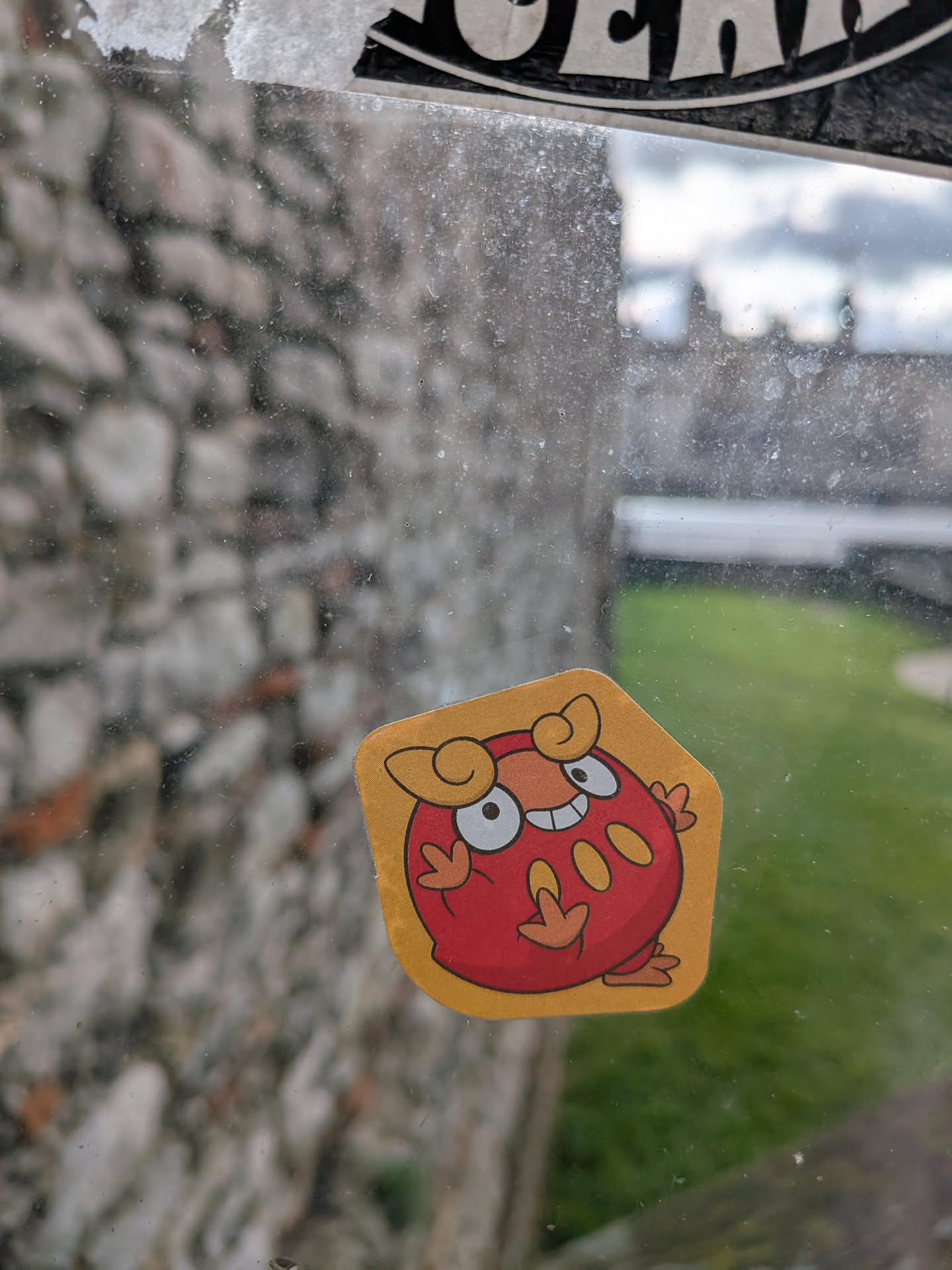 The sticker features a round, red, cartoon-style character, identifiable as Darumaka from Pokémon. It has a wide, happy smile with two visible teeth and large white eyes with black pupils looking slightly to the right. Above its eyes are two curly, yellow-orange tufts. Its body is red with three oval-shaped yellow-orange spots on its belly. It has small, three-fingered hands and feet, also in yellow-orange. The character is outlined in black, and the sticker itself has an irregular, somewhat hexagonal shape with rounded corners, bordered in a solid yellow-orange color. The sticker appears to have a slightly textured, matte finish.