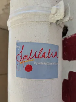 Street sticker A rectangular handmade sticker affixed to a white pole. The sticker is light blue with the text "By Lalilalia" in elegant, red cursive font. Below the name, there's a drawing of a red yarn ball and knitting needles. At the bottom right, the email address "bylalilalia@gmail.com" is printed in a smaller, simpler font. The pole is textured and weathered, with some red paint visible on the side.