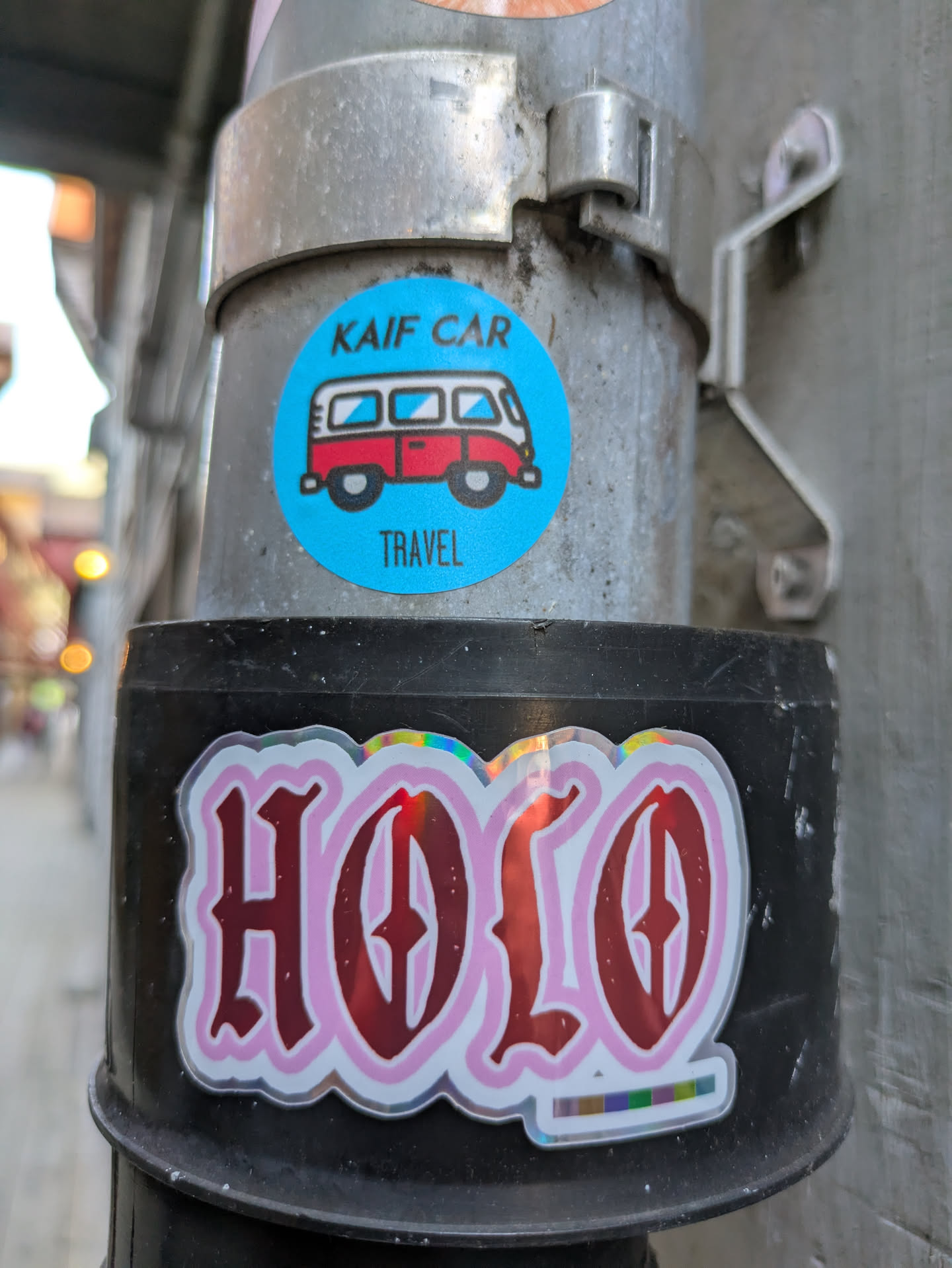 The sticker is circular with a bright blue background. It depicts a simplified, cartoonish illustration of a vintage-style red and white camper van or minibus. Above the van, the words "KAIF CAR" are printed in black, and below the van, the word "TRAVEL" is printed in black. The sticker is placed on a textured, grey metal pole, which appears to be part of a street light or utility pole, with another sticker visible below it.