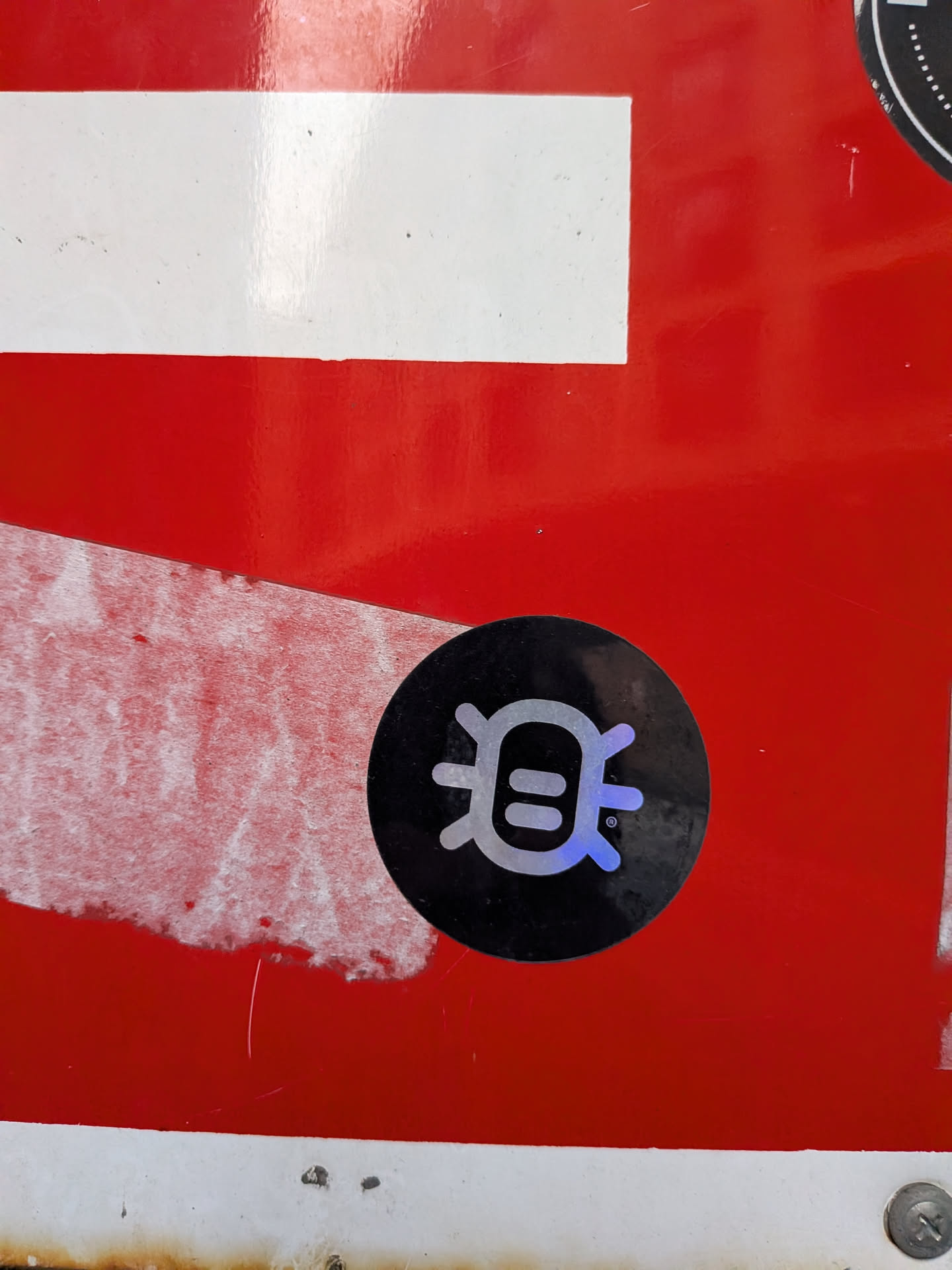 A circular sticker with a solid black background. At its center is a stylized, symmetrical symbol rendered in an iridescent or holographic material, appearing silver-white with hints of blue and purple. The symbol consists of a central vertical bar, flanked by two horizontal oval shapes, resembling eyes or a face. Curved lines extend outwards from the top and bottom, giving it an abstract, almost bug-like or alien character. A small '®' (registered trademark) symbol is visible to the right of the main icon. The sticker is applied to a vibrant red surface, which shows some signs of wear and tear.