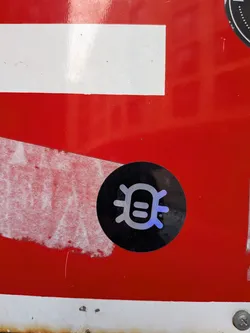 Street sticker A circular sticker with a solid black background. At its center is a stylized, symmetrical symbol rendered in an iridescent or holographic material, appearing silver-white with hints of blue and purple. The symbol consists of a central vertical bar, flanked by two horizontal oval shapes, resembling eyes or a face. Curved lines extend outwards from the top and bottom, giving it an abstract, almost bug-like or alien character. A small '®' (registered trademark) symbol is visible to the right of the main icon. The sticker is applied to a vibrant red surface, which shows some signs of wear and tear.