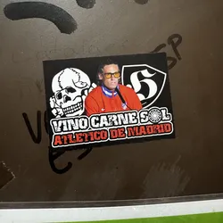 Street sticker A rectangular sticker featuring a stylized depiction of a man resembling Diego Simeone, wearing yellow sunglasses and a red tracksuit jacket with blue and white collar stripes, and a small Nike logo. To his left is a white outline of a skull, and to his right, a white stylized number '5' within a black shield with a white border. Below the central figures, the text 'VINO CARNE SOL' is displayed in distressed white letters, with a sun-like symbol between 'CARNE' and 'SOL'. Beneath this, 'ATLETICO DE MADRID' is written in distressed red letters with a white outline. The entire design is set against a black background.