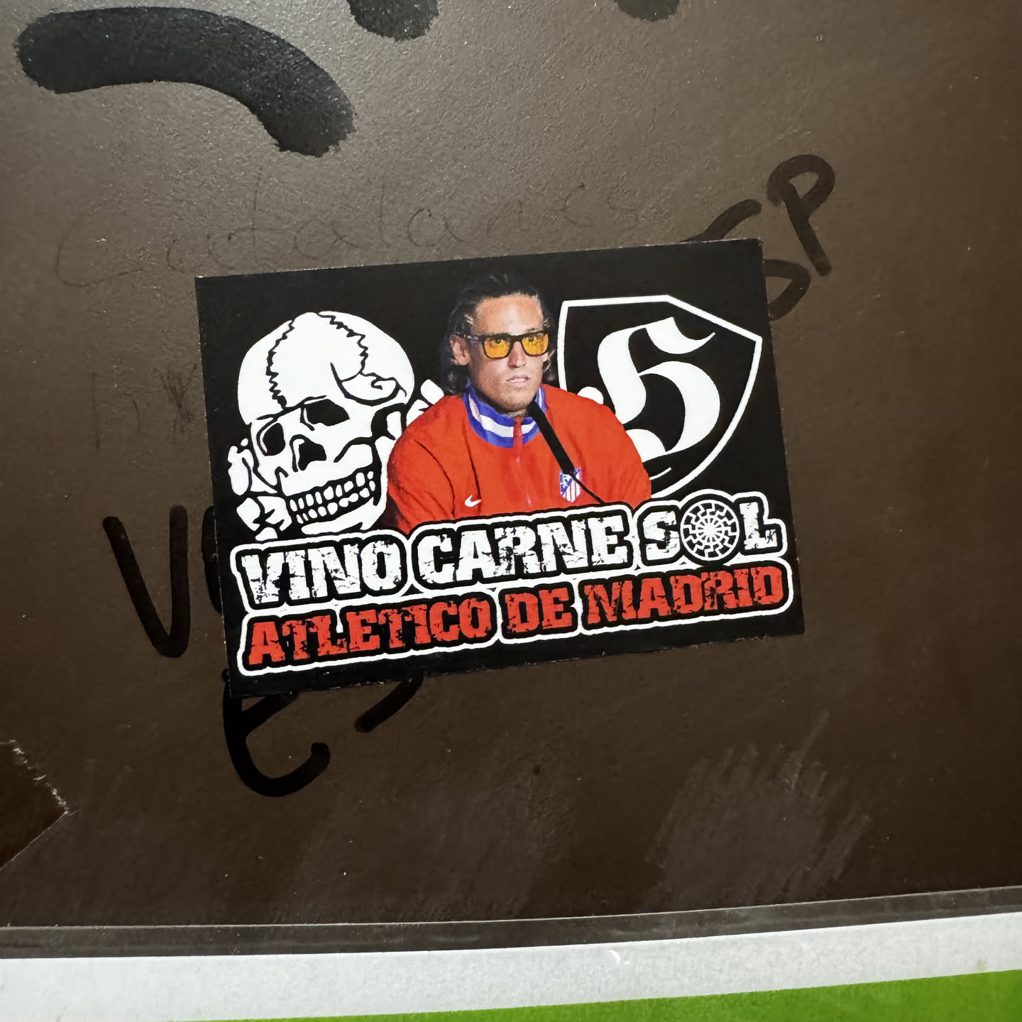 A rectangular sticker featuring a stylized depiction of a man resembling Diego Simeone, wearing yellow sunglasses and a red tracksuit jacket with blue and white collar stripes, and a small Nike logo. To his left is a white outline of a skull, and to his right, a white stylized number '5' within a black shield with a white border. Below the central figures, the text 'VINO CARNE SOL' is displayed in distressed white letters, with a sun-like symbol between 'CARNE' and 'SOL'. Beneath this, 'ATLETICO DE MADRID' is written in distressed red letters with a white outline. The entire design is set against a black background.