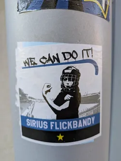 Street sticker The sticker features an illustration of a bandy player with a determined expression, raising their arm in a show of strength. The text "WE CAN DO IT!" is prominently displayed above the player. Below the illustration, the text "SIRIUS FLICKBANDY" is printed on a blue banner, followed by a yellow star. The background shows a bandy stadium.