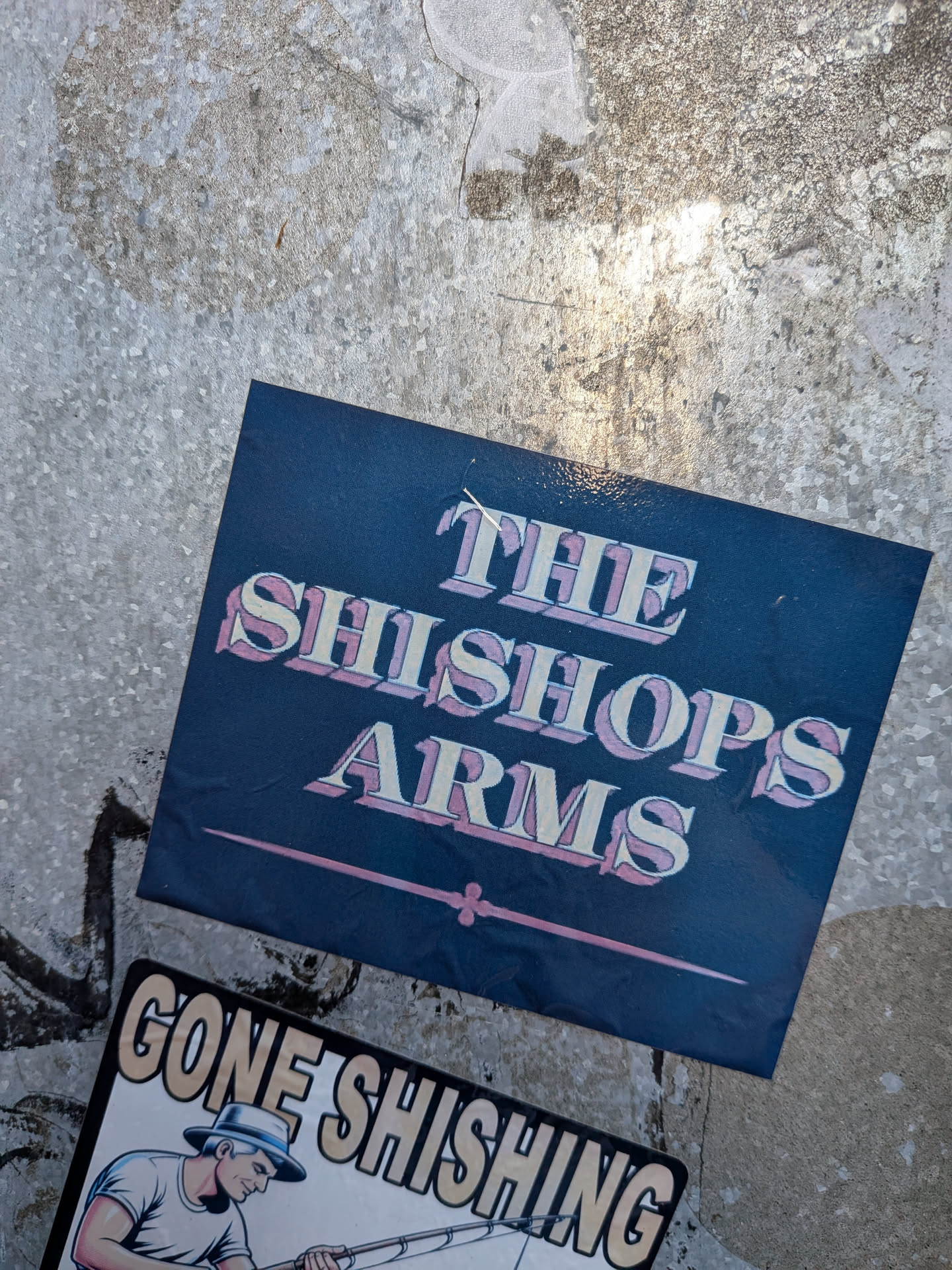 The sticker is dark blue with the text 'THE SHISHOPS ARMS' in a stylized, 3D font with cream-colored letters outlined in pink. Below the text is a thin pink line with decorative flourishes at each end. The sticker appears to be slightly creased and is adhered to a textured, metallic-looking surface. Another sticker with the text 'GONE SHISHING' and an illustration of a fisherman is partially visible below it.