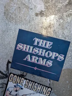Street sticker The sticker is dark blue with the text 'THE SHISHOPS ARMS' in a stylized, 3D font with cream-colored letters outlined in pink. Below the text is a thin pink line with decorative flourishes at each end. The sticker appears to be slightly creased and is adhered to a textured, metallic-looking surface. Another sticker with the text 'GONE SHISHING' and an illustration of a fisherman is partially visible below it.
