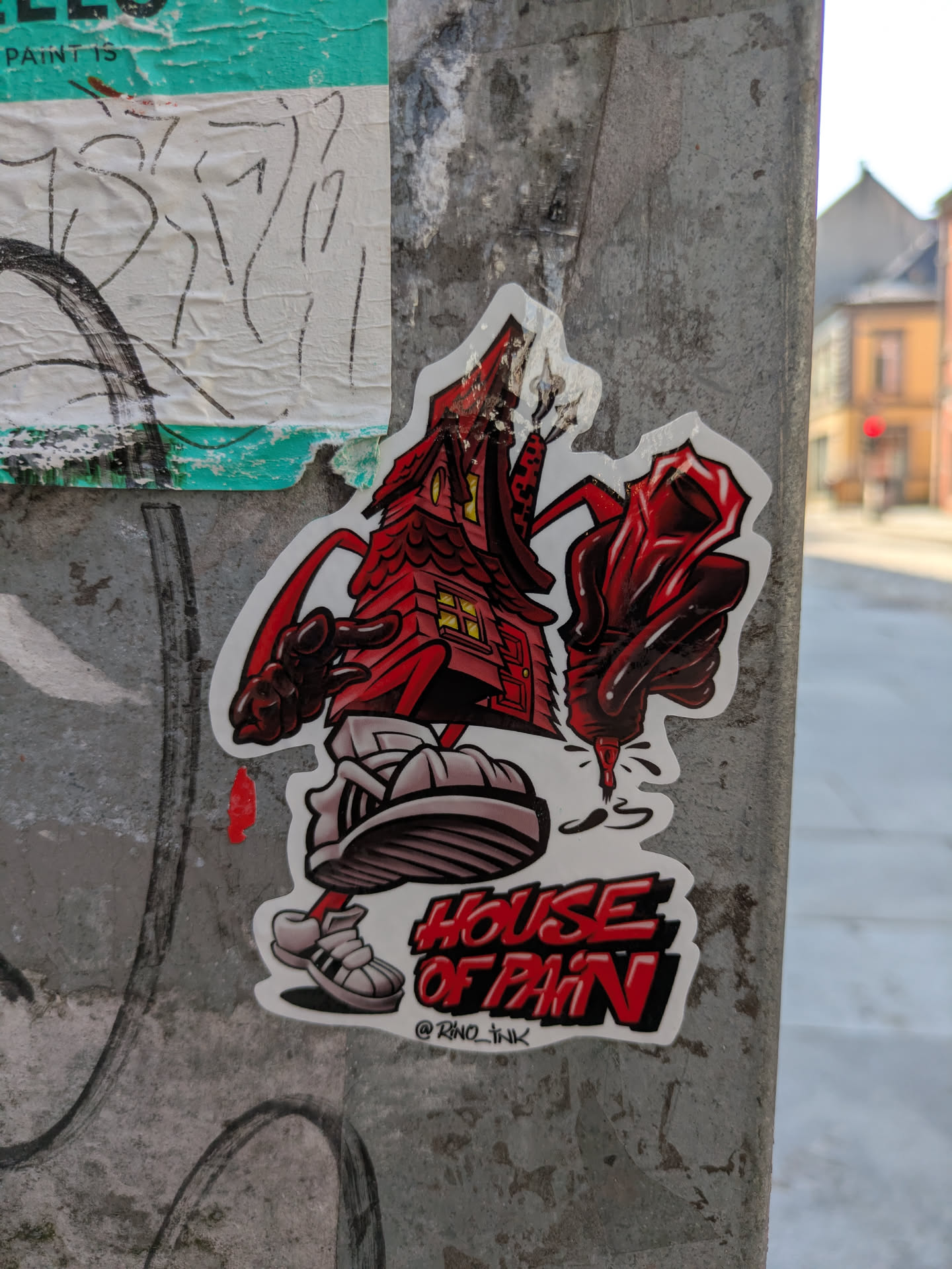 This sticker showcases a unique, cartoonish character resembling a distressed red house, complete with a jagged roof from which smoke or fire appears to emanate. The house stands on white legs adorned with large, stylized Adidas sneakers. It possesses two red arms; one is extended with an open hand, while the other grips a red and black spray paint can, dripping dark fluid onto the ground. Below the character, the bold red letters with black outlines spell out "HOUSE OF PAIN". Directly beneath this text, the artist's social media handle, "@RNO_INK", is visible. The sticker is affixed to a heavily weathered, grey utility pole, with blurred urban background details.