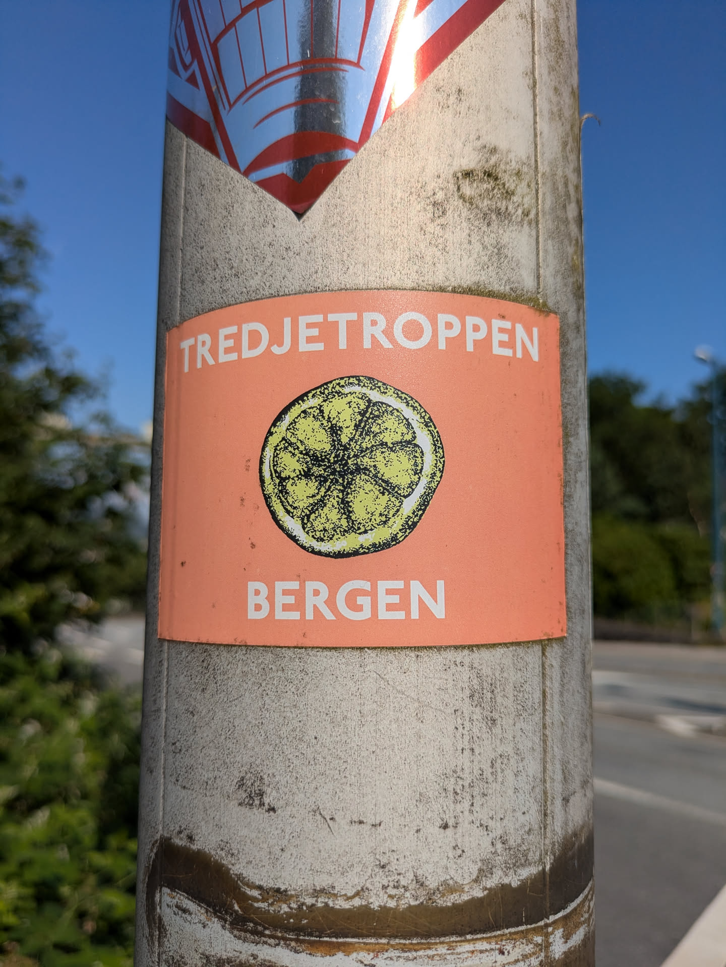 The sticker is rectangular with a prominent orange background. At the top, in white capital letters, is the word 'TREDJETROPPEN'. Below this, centrally located, is an illustration of a lemon or citrus slice, depicted with a somewhat rough, textured, and slightly speckled green-yellow interior and a white rind outline. At the bottom, also in white capital letters, is the word 'BERGEN'. The sticker is attached to a light-colored, possibly concrete or metal, pole that appears weathered and stained. Another sticker is partially visible above it.