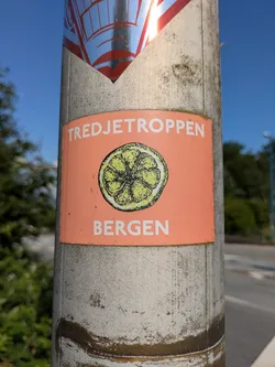 Street sticker The sticker is rectangular with a prominent orange background. At the top, in white capital letters, is the word 'TREDJETROPPEN'. Below this, centrally located, is an illustration of a lemon or citrus slice, depicted with a somewhat rough, textured, and slightly speckled green-yellow interior and a white rind outline. At the bottom, also in white capital letters, is the word 'BERGEN'. The sticker is attached to a light-colored, possibly concrete or metal, pole that appears weathered and stained. Another sticker is partially visible above it.
