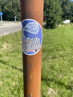 Street sticker An oval blue sticker on a rusty pole. It depicts a blonde cartoon woman with a speech bubble containing 'HA HO HE HERTHA BSC'. Below, 'Alte Dame' is written in large white cursive font.