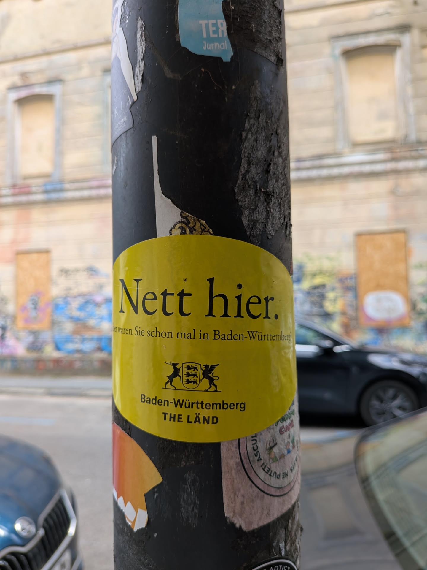 An oval yellow sticker featuring the black text 'Nett hier. Aber waren Sie schon mal in Baden-Württemberg?' alongside the state's coat of arms and the slogan 'THE LÄND'.