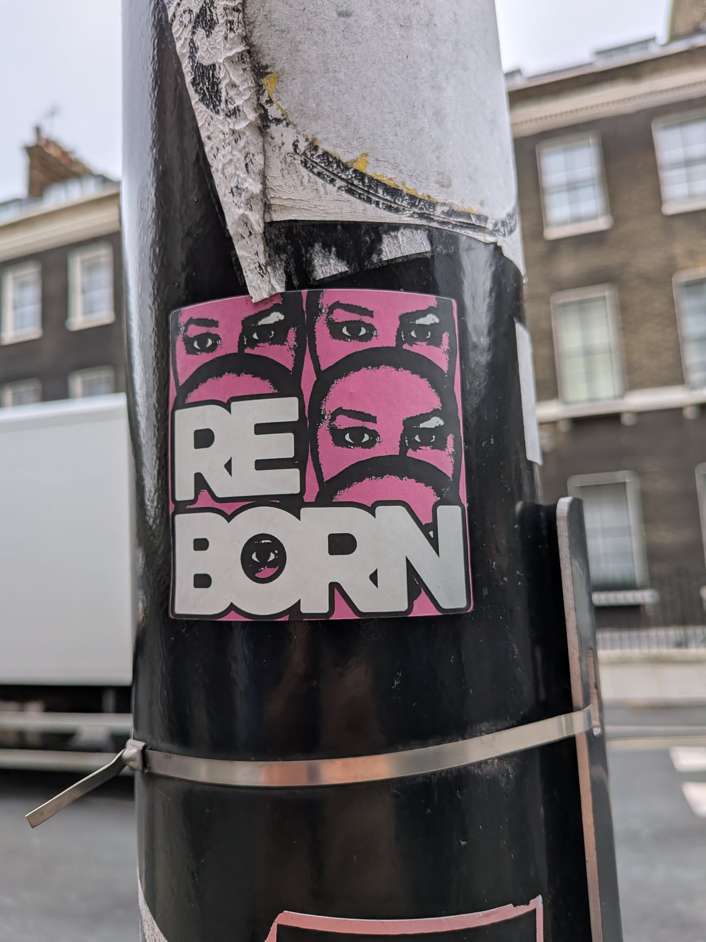 A rectangular sticker featuring a bold, graphic design. The background is a vibrant pink, upon which multiple pairs of stylized eyes are depicted in black and white, suggesting faces partially obscured or masked. The eyes are prominent, with thick black outlines and white pupils. Below these faces, the word 'REBORN' is written in large, white, blocky capital letters with a black outline. Notably, the 'O' in 'BORN' is replaced by a single, stylized eye, mirroring the eyes above. The overall aesthetic is striking and somewhat mysterious, with a strong pop art influence.