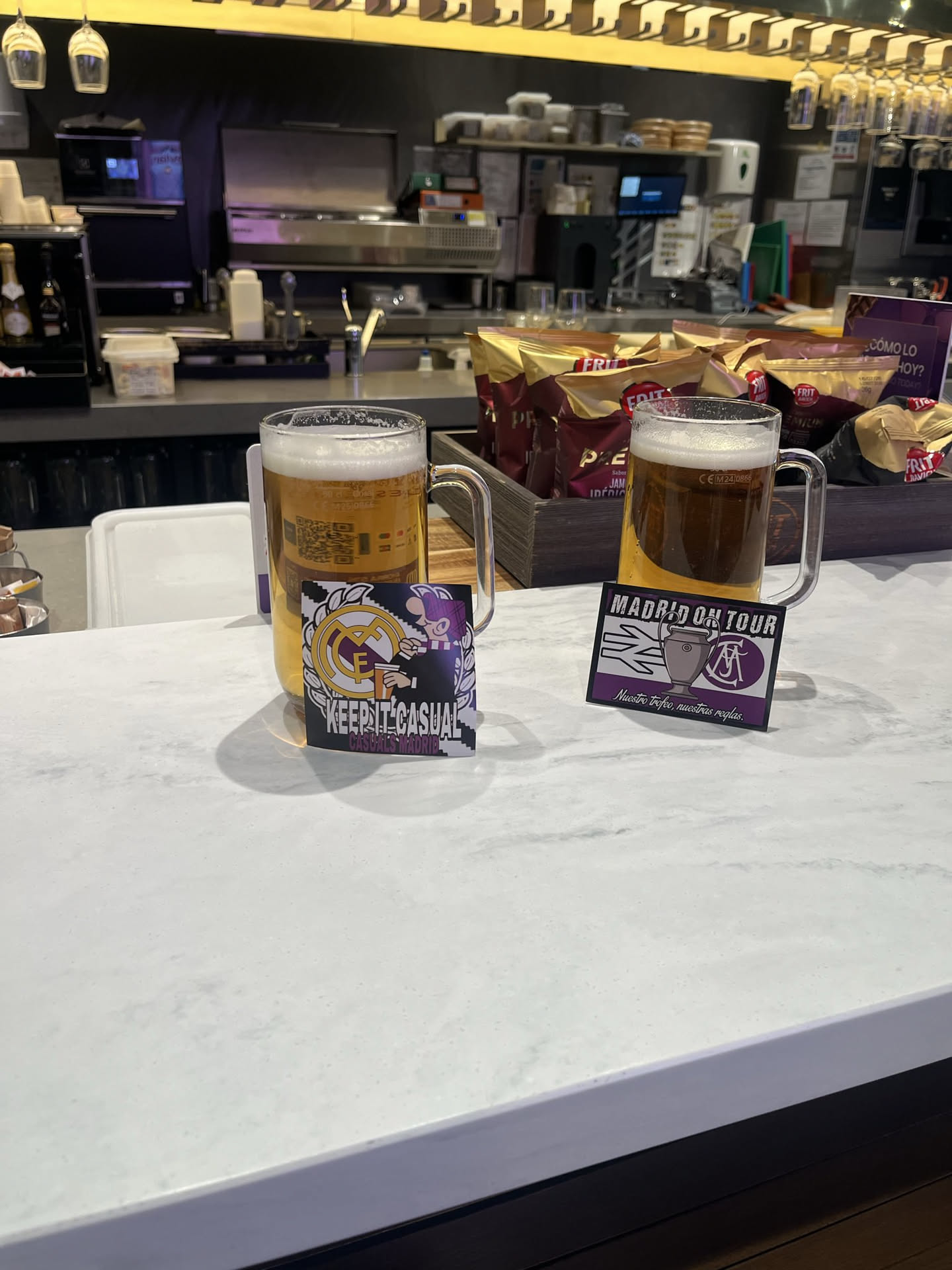 A rectangular sticker with a slightly curved bottom edge, featuring a stylized character wearing a purple beanie and a striped purple and white scarf, holding a beer mug. The iconic Real Madrid C.F. crest, in gold and purple, is visible behind the character. Bold, pixelated white text reads 'KEEP IT CASUAL' above smaller text 'CASUALS MADRID'. The design uses a palette of purple, gold, white, and black, reflecting a football fan or casuals subculture aesthetic.