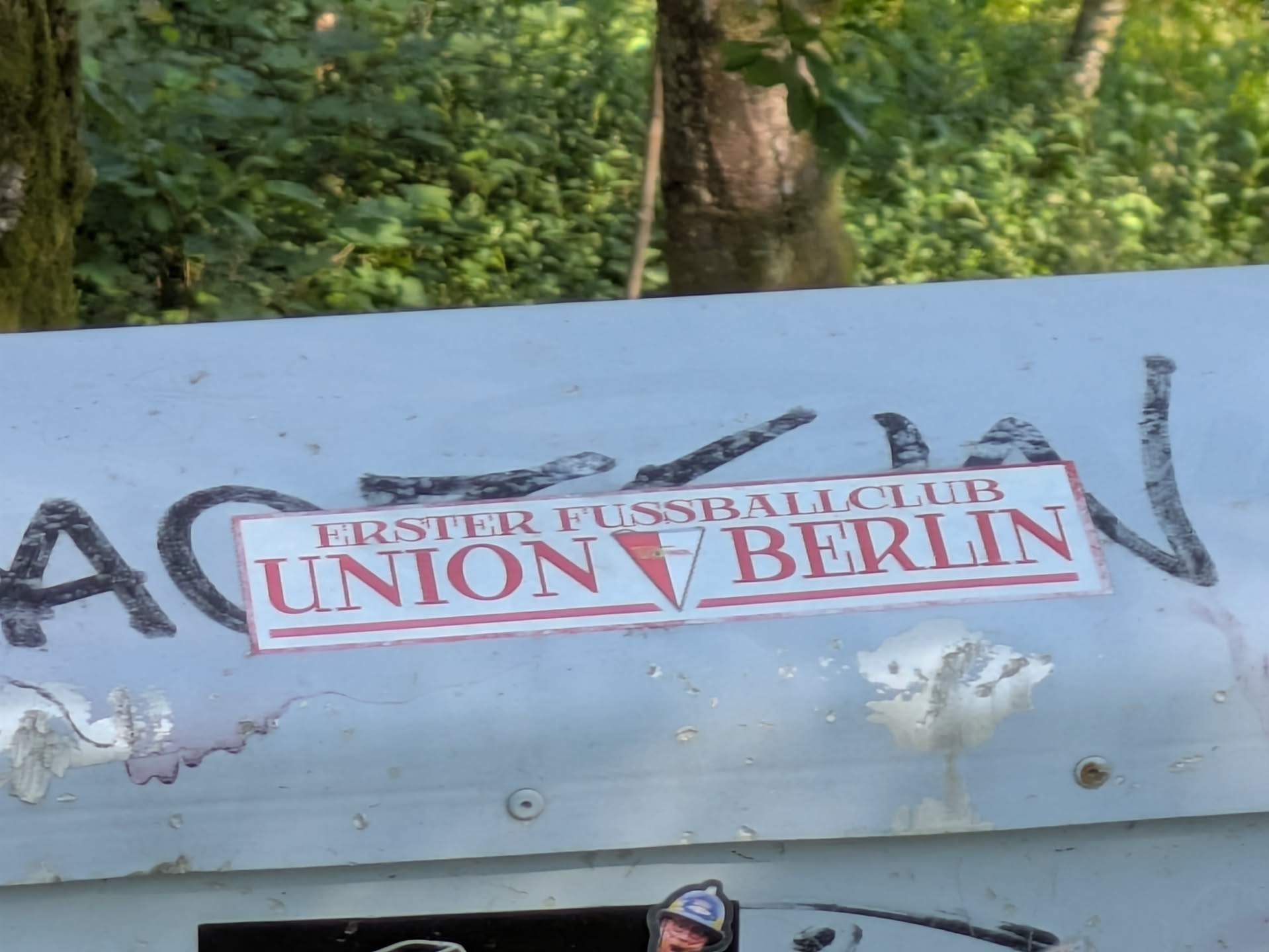 The central sticker is a white rectangle with a thin red border, featuring the text "ERSTER FUSSBALLCLUB" in smaller red font above "UNION BERLIN" in larger, bold red font. A small red triangular emblem with a 'V' shape is positioned between 'UNION' and 'BERLIN'. The sticker is placed on a light grey or white surface that shows signs of wear, discoloration, and black graffiti marks. In the background, green foliage and trees are visible, suggesting an outdoor setting.