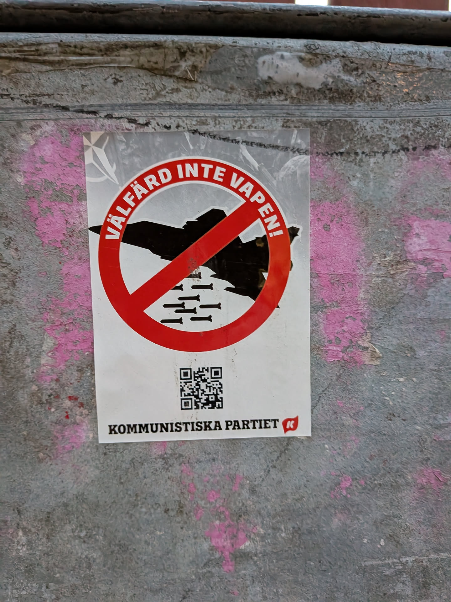 The sticker features a prominent red circle with a diagonal line through it, indicating prohibition. Inside the circle, a silhouette of a jet fighter is depicted, with several bombs falling from it. The text 'VÄLFÄRD INTE VAPEN!' is written in white capital letters along the curve of the red prohibition symbol. Below this, a QR code is visible, and further down, the text 'KOMMUNISTISKA PARTIET' is printed in black, accompanied by a small red logo with a white 'K'. The sticker is white with red and black elements, and the background of the sticker appears to be a light grey with a faint NATO symbol visible in the upper left corner.