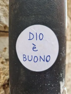 Street sticker A round white sticker with blue handwriting on a dark gray metallic surface. The text on the sticker says "DIO è BUONO", which is Italian for "God is good".