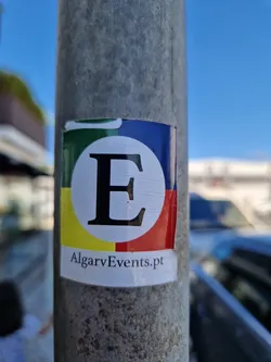 Street sticker A sticker featuring a large letter 'E' in a circle with a color block design and the text 'AlgarveEvents.pt' below. The sticker is affixed to a metal pole.