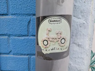 The sticker is placed on a gray pole, partially torn at the bottom. It features the text "BikeDesuro" at the top and a bicycle design made of cork pieces. The text "La vie in bike La vie in bike" is written at the bottom of the design.