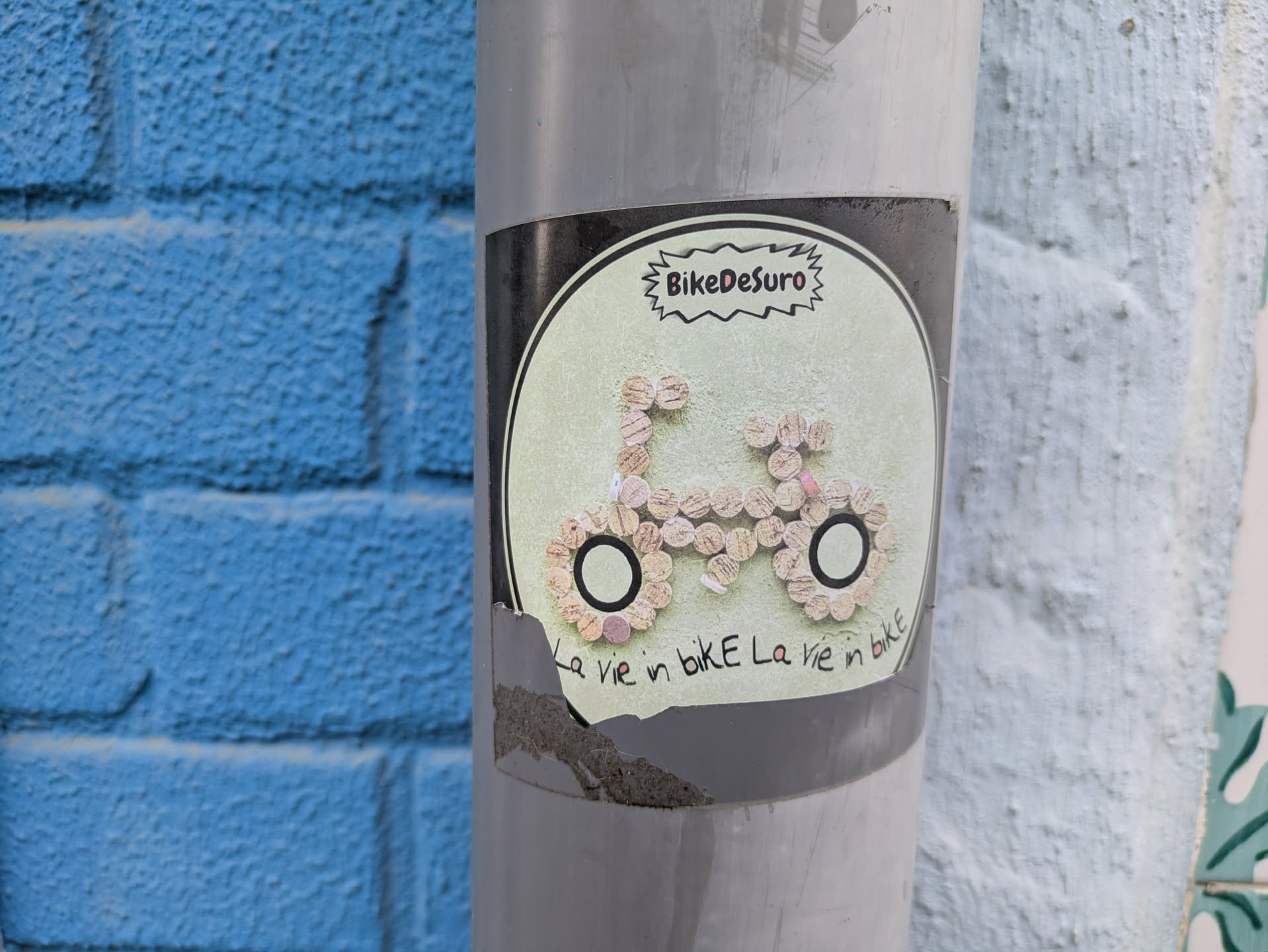Street sticker The sticker is placed on a gray pole, partially torn at the bottom. It features the text "BikeDesuro" at the top and a bicycle design made of cork pieces. The text "La vie in bike La vie in bike" is written at the bottom of the design.