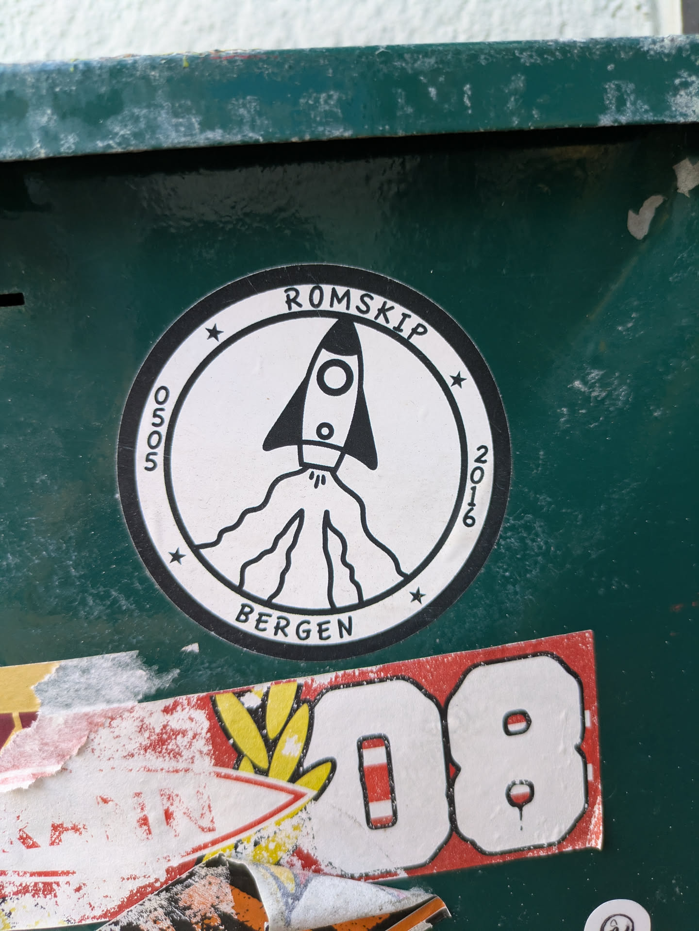 This is a circular black and white sticker affixed to a dark green, somewhat scuffed surface. The central design depicts a stylized rocket ship in mid-launch, with its exhaust plumes extending downwards. The rocket is enclosed within a circular border. Along the top of the circle, the word 'ROMSKIP' is written, and along the bottom, 'BERGEN' is visible. On the left side of the circle, '0505' is written vertically, and on the right side, '2016' is written vertically. Small stars are placed at intervals along the inner rim of the circular border. The sticker has a clean, minimalist aesthetic.