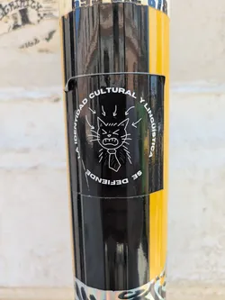 The black and white sticker features a drawing of an angry cat wearing a tie. The text surrounding the cat reads "LA IDENTIDAD CULTURAL Y LINGUISTICA SE DEFIENDE".