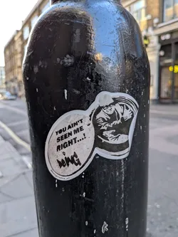 Street sticker The sticker is an irregularly shaped, monochrome design, primarily black and white with a thin white border. It features a stylized, somewhat menacing character on the right, depicted in a cap and possibly a jacket, with an intense expression and arms held close to the body. To the left of the character, a speech bubble contains the main text. Below the main text, there's a smaller, stylized signature or tag. The sticker is applied to a textured, dark pole, showing some signs of wear and slight peeling at the edges.