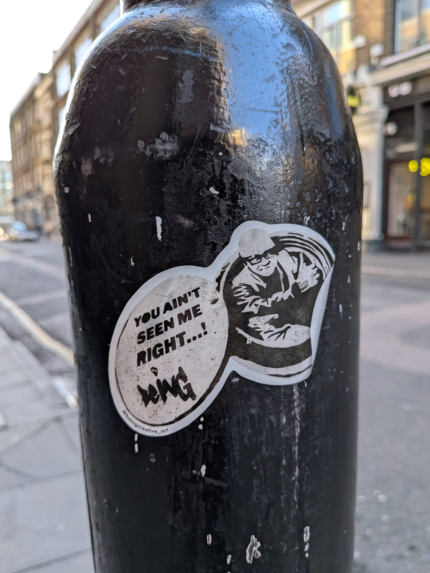 The sticker is an irregularly shaped, monochrome design, primarily black and white with a thin white border. It features a stylized, somewhat menacing character on the right, depicted in a cap and possibly a jacket, with an intense expression and arms held close to the body. To the left of the character, a speech bubble contains the main text. Below the main text, there's a smaller, stylized signature or tag. The sticker is applied to a textured, dark pole, showing some signs of wear and slight peeling at the edges.