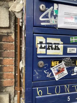 Street sticker The central sticker is a small, square paper with a white background. It features a photograph in the center which appears to depict a somber or difficult scene, with several people gathered around. Around the photograph, there is text in Dutch. The top line, below the image, reads 'DIT IS GEEN MAALTIJD'. The line below that reads 'DIT IS MOORD' and then 'STOP DEZE HOLOCAUST'. The final line reads 'IEDEREEN HEEFT RECHT OP LEVEN'. The sticker appears slightly creased.