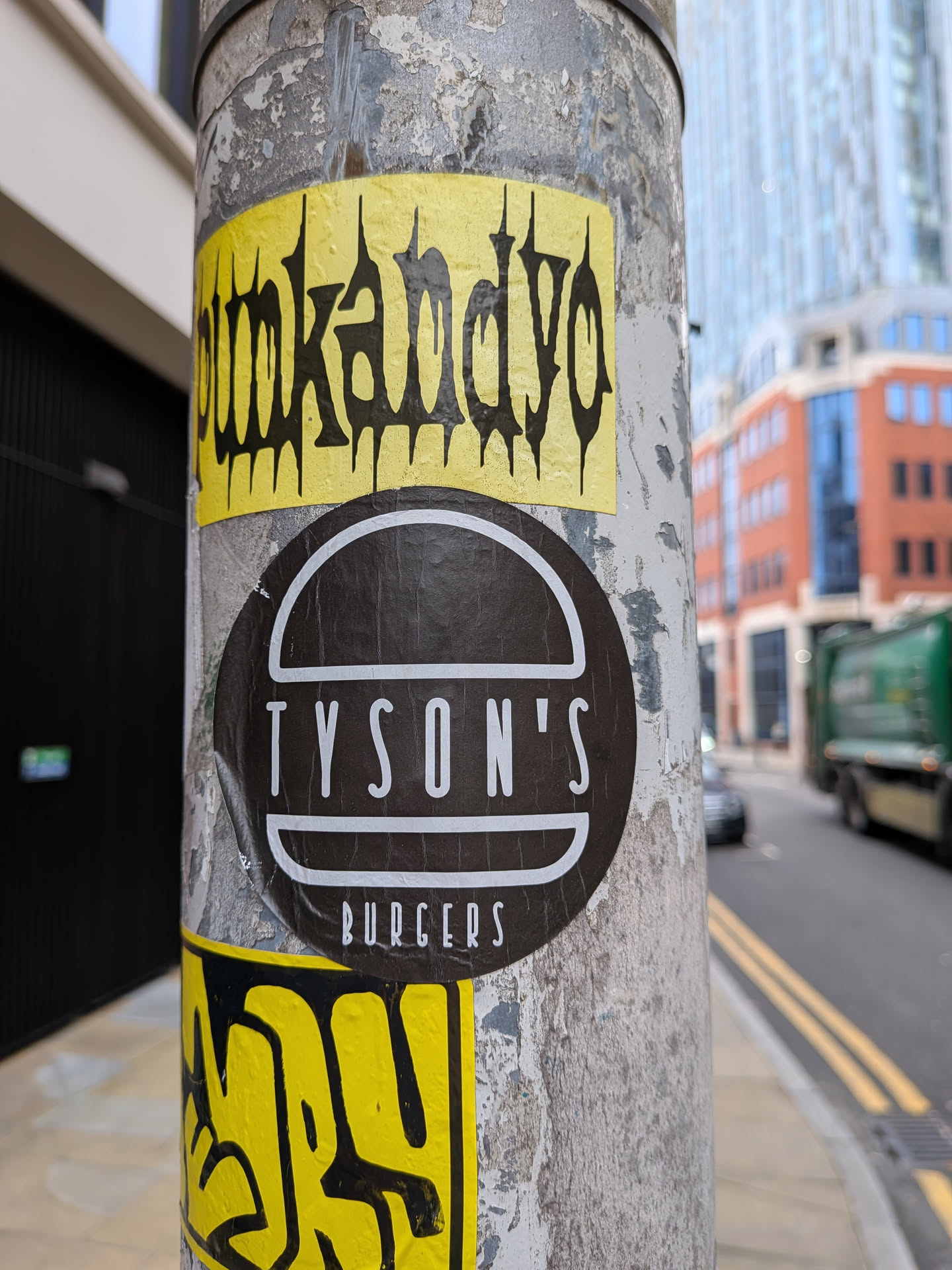 The central sticker is circular with a dark brown or black background. It prominently displays the word 'TYSON'S' in large, white, sans-serif capital letters. Below 'TYSON'S', a thinner white horizontal line separates it from the word 'BURGERS', which is also in white, sans-serif capital letters but smaller in size. Above 'TYSON'S', a thick white semi-circular line forms an arc, completing the logo's design. The sticker shows signs of wear, with slight wrinkles and peeling around its edges, indicating it's a physical sticker adhered to the pole.