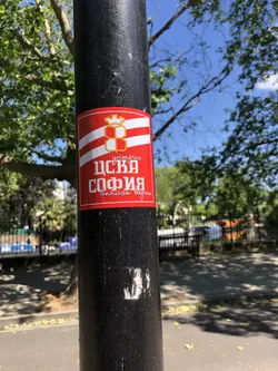 Street sticker A rectangular sticker with a prominent red background, featuring a white and red striped pattern across its upper half. Centered on the stripes is a golden crest, resembling a shield with a cross design, topped by a crown or castle turret. Below the stripes, on the solid red background, is white Cyrillic text. The main text 'ЦСКА СОФИЯ' is large and bold, with smaller, cursive text 'Шитрас' above it and 'винаги верни' below. The sticker has a slight sheen and appears to be adhered to a black metal pole.
