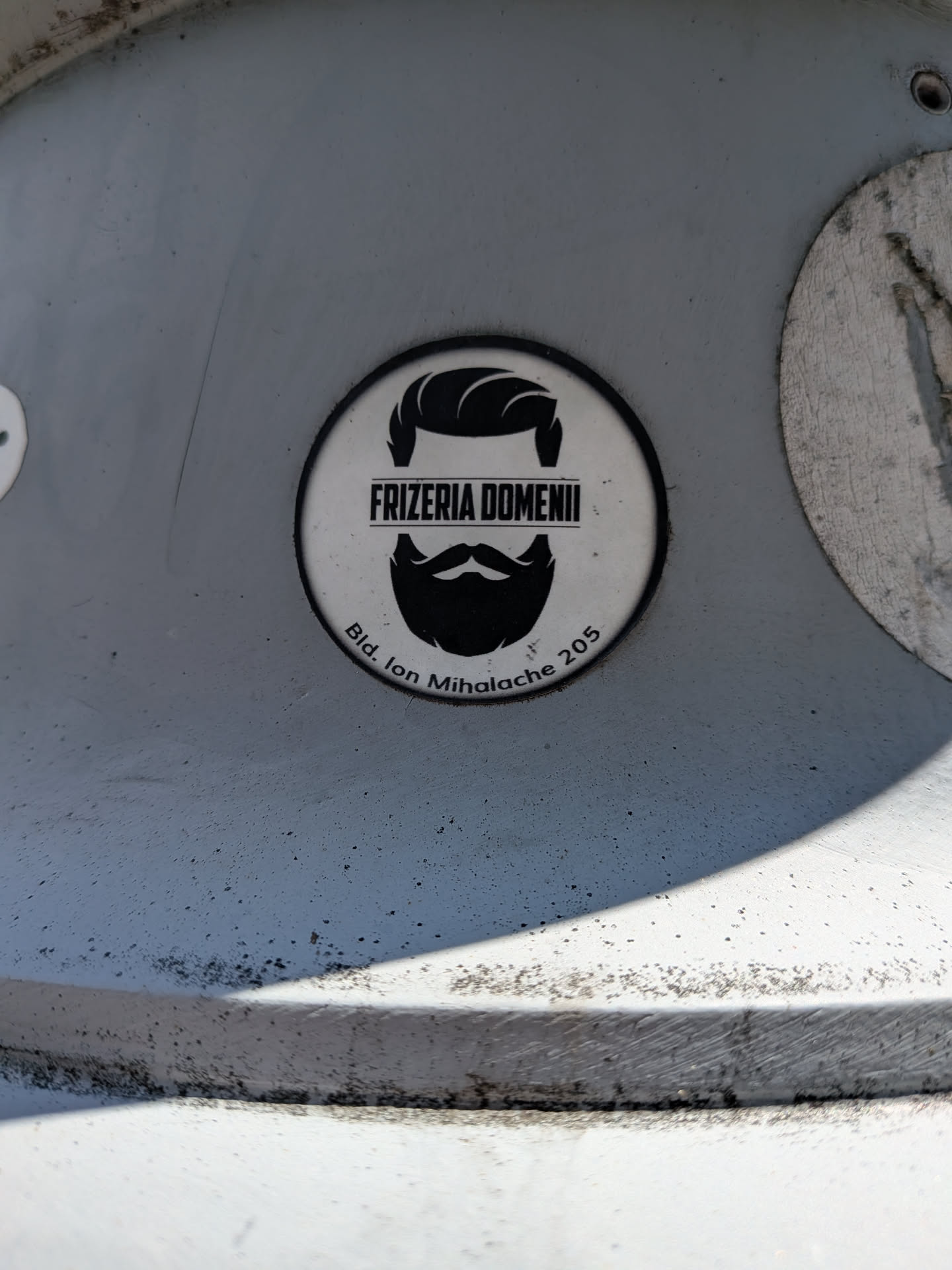 A round white sticker with a black border, featuring a black silhouette of a styled haircut, mustache, and beard. The text 'FRIZERIA DOMENII' is printed in the center, with the address 'Bld. Ion Mihalache 205' curved along the bottom.