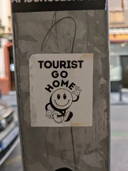 This sticker features a smiling cartoon figure with the text 'TOURIST GO HOME' above it. The sticker is placed on a gray metal pole.
