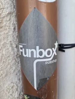 Street sticker A diamond-shaped sticker with the Funbox skateshop logo. The logo is predominantly white with the text "Funbox" prominently displayed, with "skateshop" written in smaller text underneath. The background of the sticker is dark gray. The sticker is affixed to a brown cylindrical surface, possibly a pole or pipe.