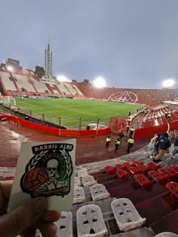 Street sticker The sticker features a central circular emblem with a stylized white skull wearing a dark cap, set against a dark green background. Below the skull, a white sneaker with red, white, and green stripes is depicted, alongside an orange-brown football. The entire circular design is framed by a green laurel wreath. Above the wreath, in an arch, is the text 'BARRIO ALBO' in bold white letters with a black outline. Below the skull, within the green circle, 'LA SULTANA' is written above the skull and 'LA BANDA' is written below the skull. The sticker has a black outline around its irregular shape, which is roughly rectangular with rounded corners, and is held against the backdrop of a football stadium.