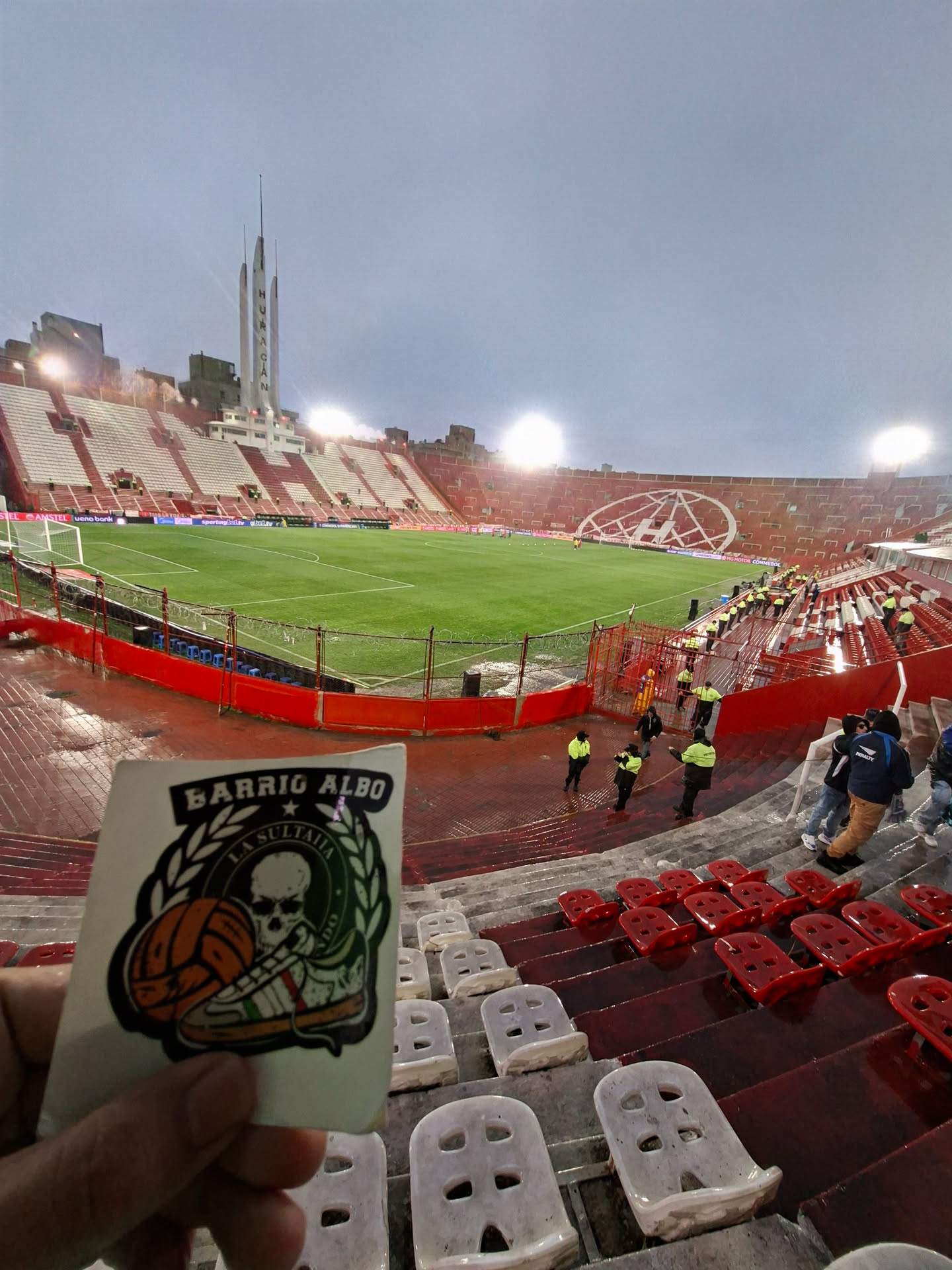 The sticker features a central circular emblem with a stylized white skull wearing a dark cap, set against a dark green background. Below the skull, a white sneaker with red, white, and green stripes is depicted, alongside an orange-brown football. The entire circular design is framed by a green laurel wreath. Above the wreath, in an arch, is the text 'BARRIO ALBO' in bold white letters with a black outline. Below the skull, within the green circle, 'LA SULTANA' is written above the skull and 'LA BANDA' is written below the skull. The sticker has a black outline around its irregular shape, which is roughly rectangular with rounded corners, and is held against the backdrop of a football stadium.