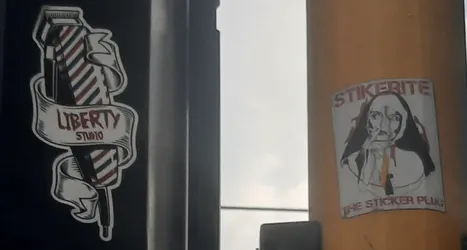 Street sticker The sticker depicts a black and white stylized illustration of a person's face, possibly a man, with long dark hair or a hood. The hands are clasped near the chin, suggesting contemplation or a similar pose. There are subtle red and orange color accents within the illustration, particularly around the face and on what appears to be a tie or garment. Above the face, the word 'STIKERITE' is written in a bold, capitalized font. Below the illustration, the phrase 'THE STICKER PLUG' is written.