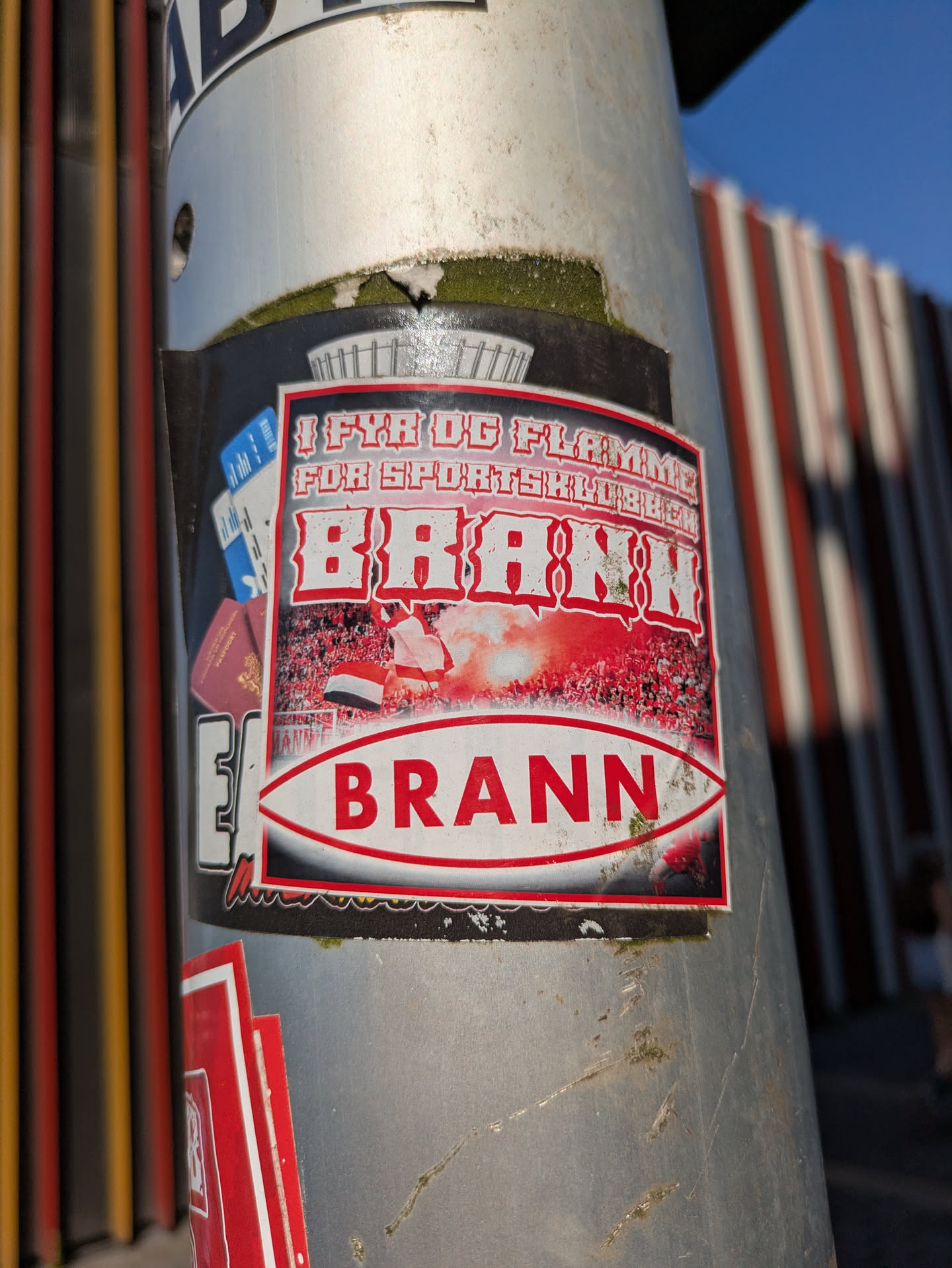 The sticker is centered on a grey metal pole, showing signs of wear with some tearing and dirt. The main design is a red, white, and black rectangular sticker with a prominent football theme. At the top, in a stylized font, is the phrase "I FYR OG FLAMME FOR SPORTSKLUBBEN BRANN". Below this, there's an image depicting a packed stadium crowd with a large amount of red smoke or flares, and a flag visible. Below the stadium scene, in a red banner with white text, the word "BRANN" is clearly written. To the left of the main sticker, partially visible behind it, are graphics resembling what might be a passport and some tickets, suggesting travel or attendance for matches. The overall impression is that of a passionate supporter's emblem for a sports team.