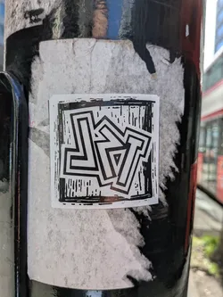 Street sticker A square, black and white sticker featuring highly stylized, interlocking block letters. The letters, which appear to spell 'LYT', are outlined in black with white interiors, creating a strong contrast. The background within the sticker is a textured black with white striations, reminiscent of a woodcut or linocut print, giving it a raw, artistic feel. The overall design is graphic and angular, with the letters forming a cohesive, abstract pattern. The sticker is applied to a dark, possibly metal pole, over what appears to be older, faded white sticker residue.