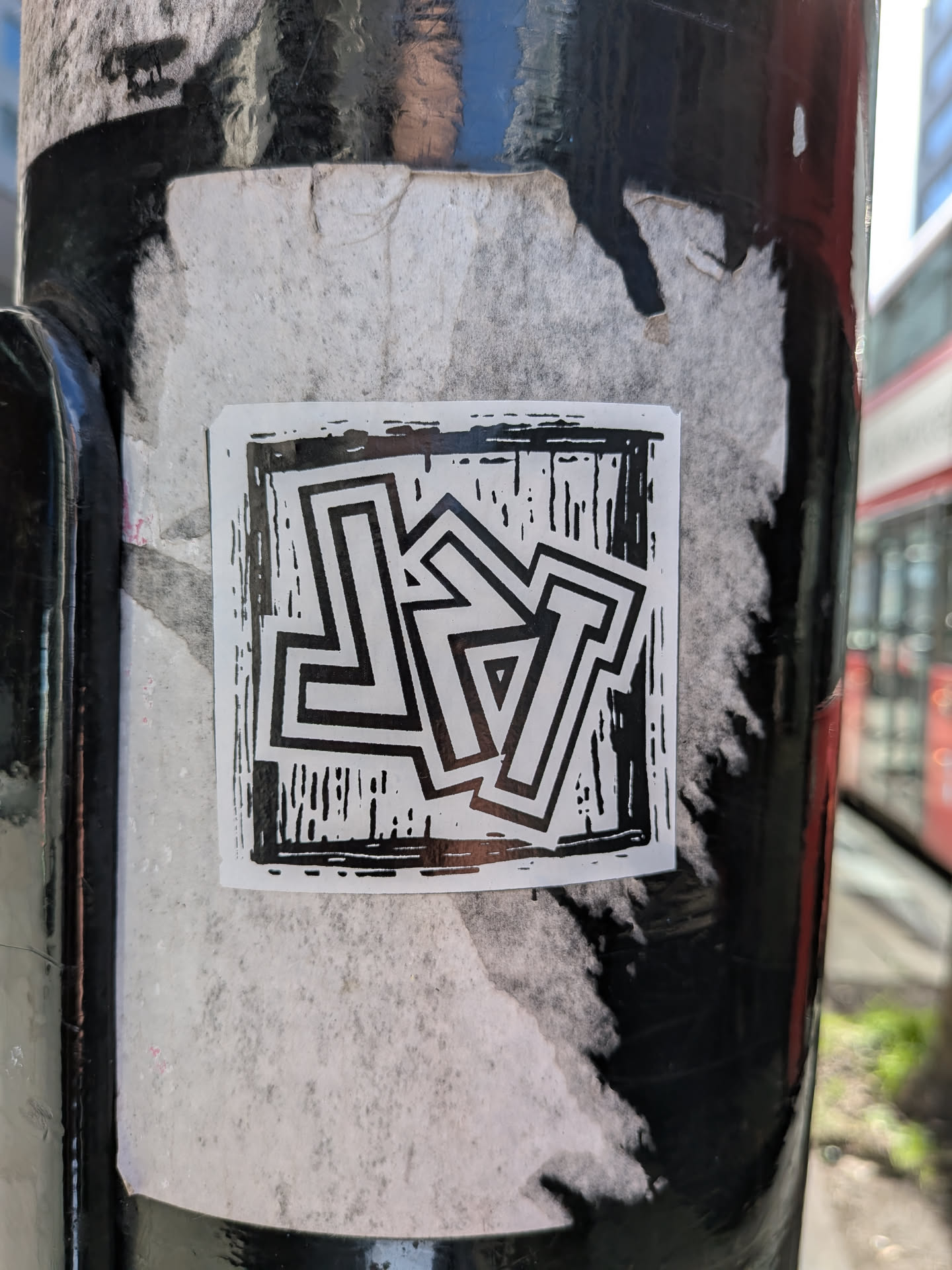 A square, black and white sticker featuring highly stylized, interlocking block letters. The letters, which appear to spell 'LYT', are outlined in black with white interiors, creating a strong contrast. The background within the sticker is a textured black with white striations, reminiscent of a woodcut or linocut print, giving it a raw, artistic feel. The overall design is graphic and angular, with the letters forming a cohesive, abstract pattern. The sticker is applied to a dark, possibly metal pole, over what appears to be older, faded white sticker residue.