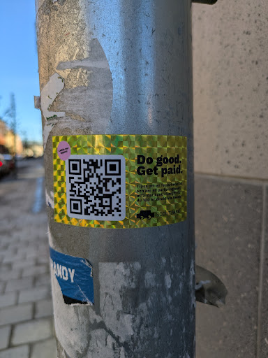 The sticker features a holographic background and black text, along with a QR code for more information. It's attached to a metal pole.