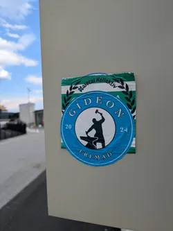 Street sticker The central sticker is round and predominantly blue with a white inner circle. Inside this circle is a black silhouette of a person, likely a blacksmith, striking an anvil with a hammer. Around the outer blue ring, the word "GIDEON" is written at the top, "FREMAD" at the bottom, and the numbers "20" and "24" are placed on the left and right sides respectively, suggesting a year. Partially visible behind this blue sticker is a rectangular sticker with horizontal green and white stripes and a black laurel wreath design, with text "Haseta Kollektiv" visible.