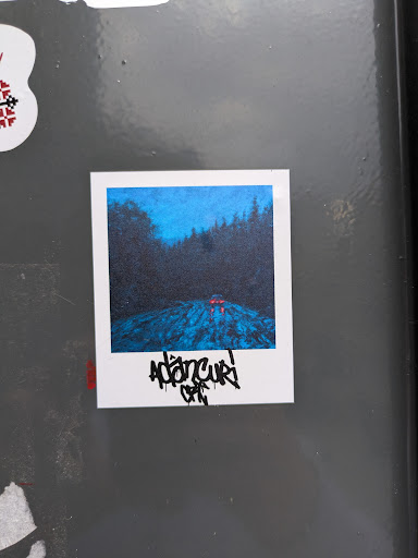 The sticker features a polaroid-style photo of a forest. It appears to be nighttime with a road and trees and two red lights on the road. Below the photo, there is some handwritten text.