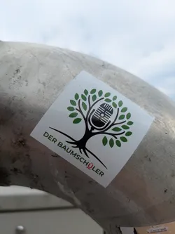 Street sticker The sticker showcases a stylized black tree with a full canopy of vibrant green leaves. Instead of a traditional trunk or fruit, an old-fashioned black microphone is centrally embedded within the tree's branches, symbolizing a connection between nature and audio media. Below the tree, the text 'DER BAUMSCHÜLER' is printed, predominantly in green, with the umlaut 'Ü' in 'SCHÜLER' highlighted in red. The sticker is square with slightly rounded corners and is affixed to a textured, metallic pipe or rail.
