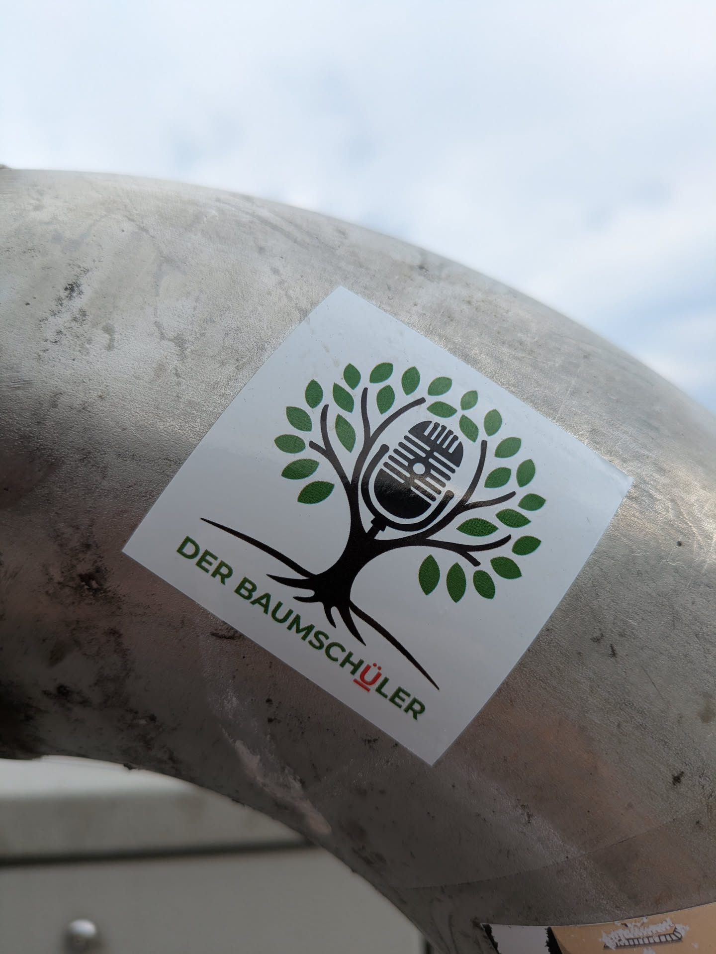 The sticker showcases a stylized black tree with a full canopy of vibrant green leaves. Instead of a traditional trunk or fruit, an old-fashioned black microphone is centrally embedded within the tree's branches, symbolizing a connection between nature and audio media. Below the tree, the text 'DER BAUMSCHÜLER' is printed, predominantly in green, with the umlaut 'Ü' in 'SCHÜLER' highlighted in red. The sticker is square with slightly rounded corners and is affixed to a textured, metallic pipe or rail.