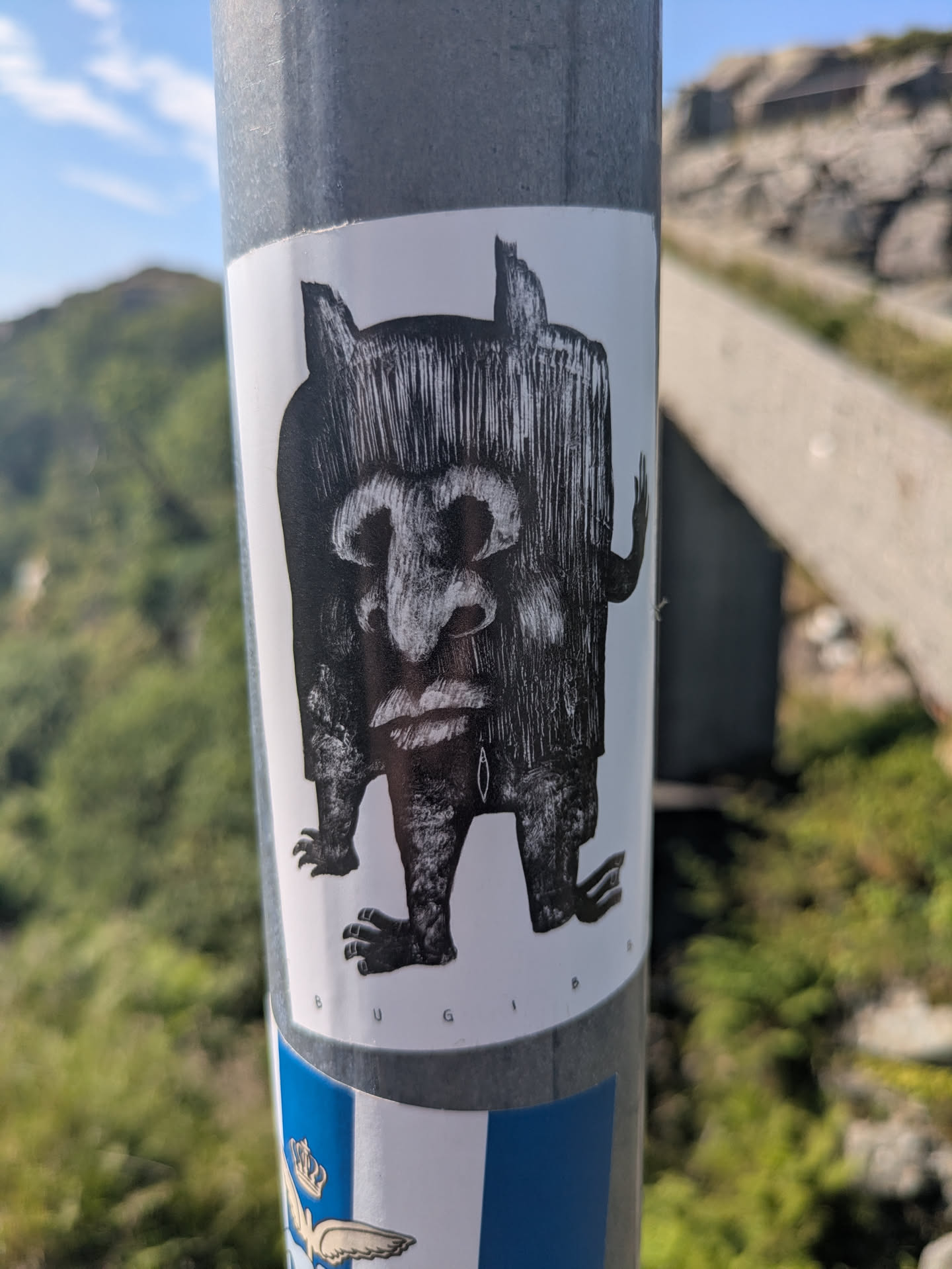 The sticker features a monochrome illustration of a squat, robust creature with a distinctive, somewhat primitive appearance. Its body is covered in a dense pattern of vertical lines, resembling thick fur or hair, which extends upwards to form ear-like protrusions. The face has a large, fleshy nose, full lips, and what appears to be a single, vertical line or mark below the left eye. It has small, clawed hands and feet, giving it a rugged, ancient, mythical feel. The background shows a blurred natural environment with greenery and rocks.