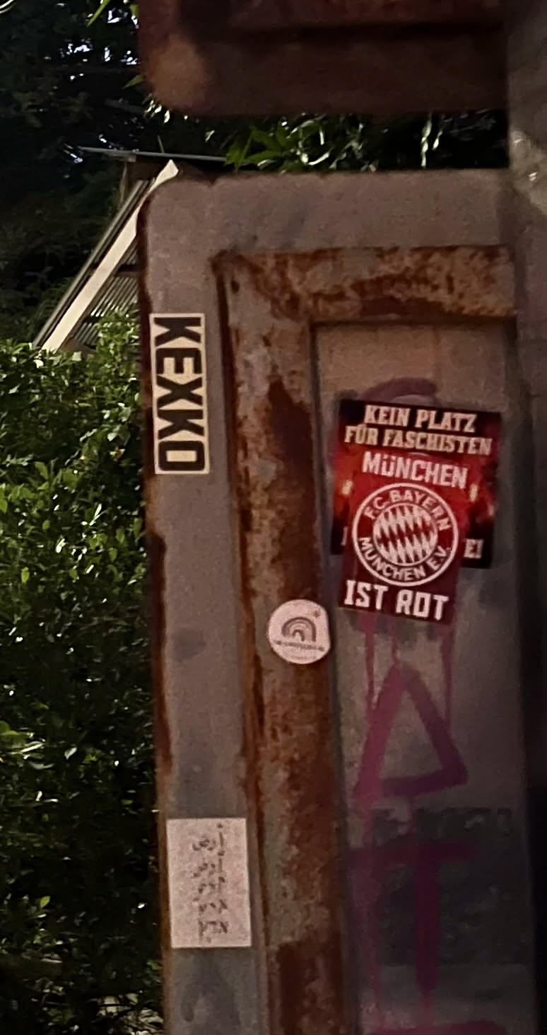 The sticker is rectangular, predominantly red with white text and a white border. At the top, in bold white capital letters, it reads "KEIN PLATZ FÜR FASCHISTEN" (No place for fascists). Below this, also in white, is "MÜNCHEN" (Munich). The central and most prominent element is the circular FC Bayern München logo, rendered in white with red details, including the club's name and the Bavarian flag pattern. Below the logo, in white capital letters, it states "IST ROT" (Is Red). The background of the sticker is a vibrant red, possibly with some subtle texture or light effects, giving it an energetic appearance.