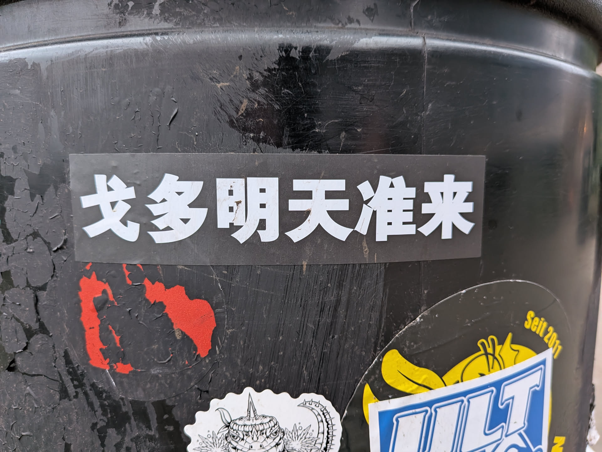 A rectangular sticker with a matte black background featuring five bold white Chinese characters. The characters are evenly spaced and appear to be a standard, clear font. The sticker is applied to a heavily worn, scratched, and peeling black surface, which appears to be a metal or plastic bin or pole. The edges of the sticker are clean, suggesting a die-cut design.