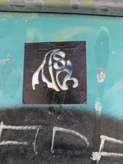 This square sticker is adhered to a surface, likely outdoors, and features an abstract symbol. The design is rendered in what appears to be chrome, with a black background. The symbol is difficult to discern, as it is not a standard letterform or easily recognizable shape. The sticker has some wear and tear, with some imperfections visible on the surface.